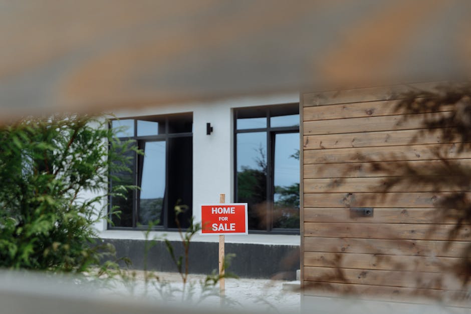 A contemporary home with a 'For Sale' sign in the front yard, framed by greenery